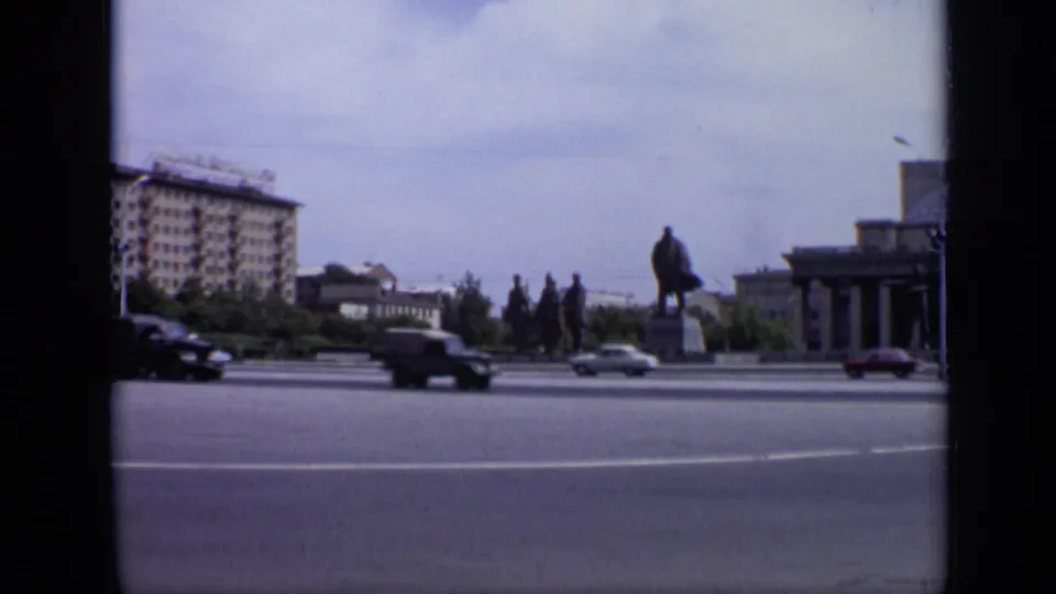 1971 Moscow street frame showing classic cars crossing the foreground with a statue, apartment block, trees, and part of a columned building behind.