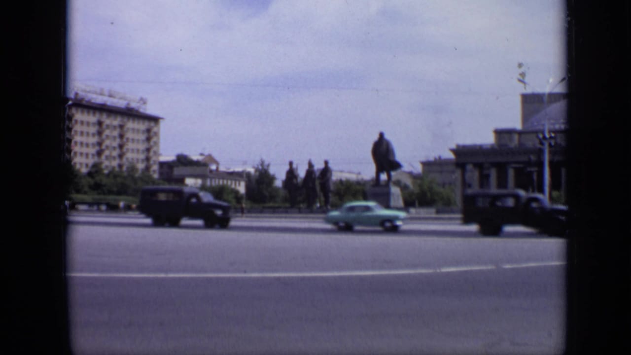 Wide street view with Lenin monument, passing cars, large building, trees, and pale sky.