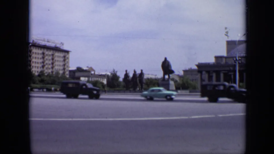 1971 Soviet city square view with a Lenin monument, passing sedans, a large civic building, trees, and broad roadway.