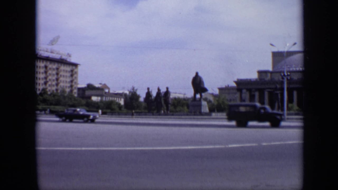 Lenin monument beside a wide road with dark cars, distant pedestrians, trees, and civic buildings.