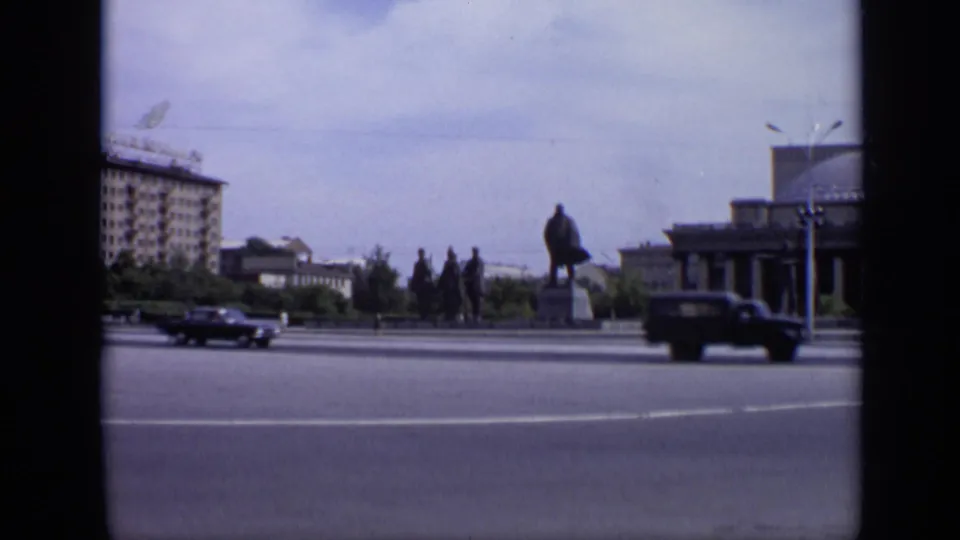 1971 Moscow street view with the Lenin monument, dark sedans moving through the intersection, distant pedestrians, trees, and civic buildings.