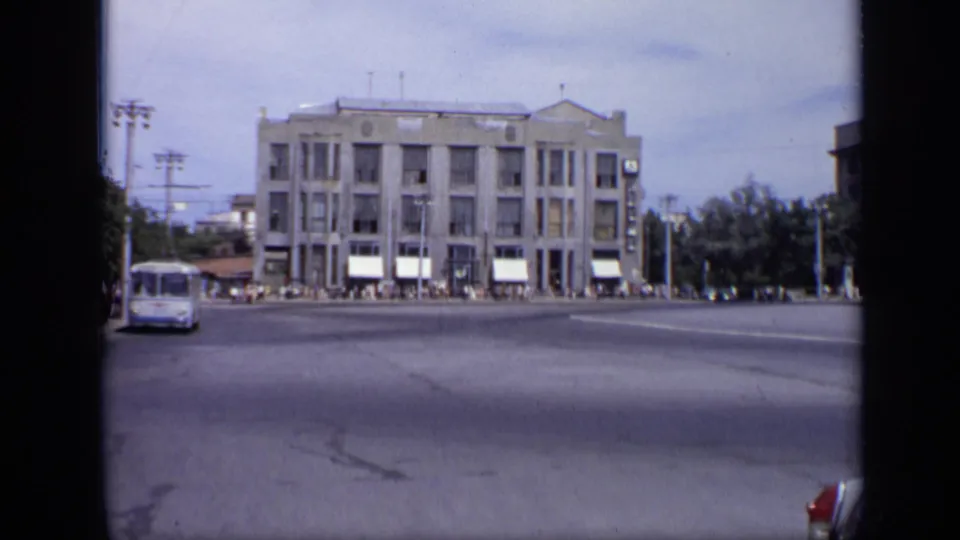 1971 Soviet city intersection with a large civic building, white awnings, bus at left, pedestrians along the facade, poles, and trees.