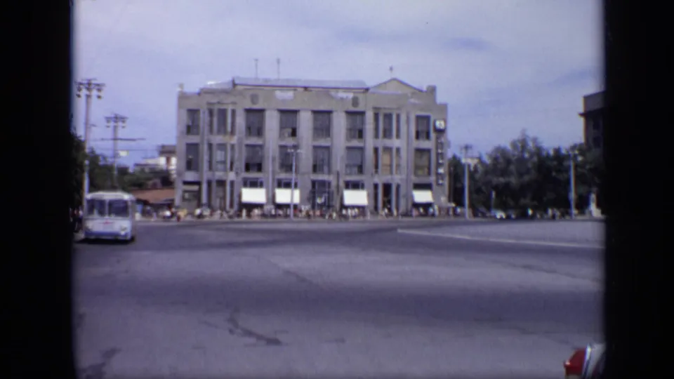 1971 Soviet-era city view facing a large civic building across a wide road, with a bus, white awnings, pedestrians, trees, and utility poles.