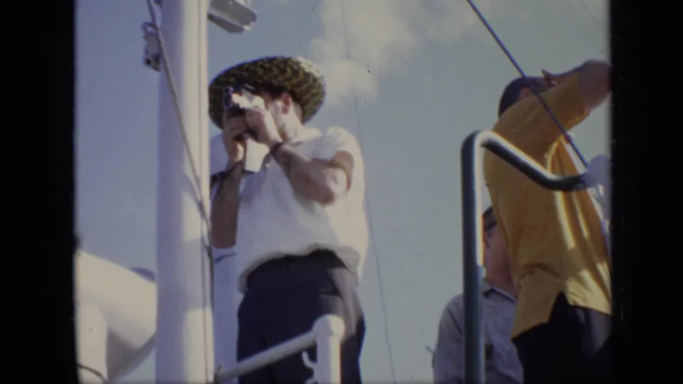 1971 8mm home-movie frame of passengers on a ship deck, with a man in a patterned hat raising a camera to his face.