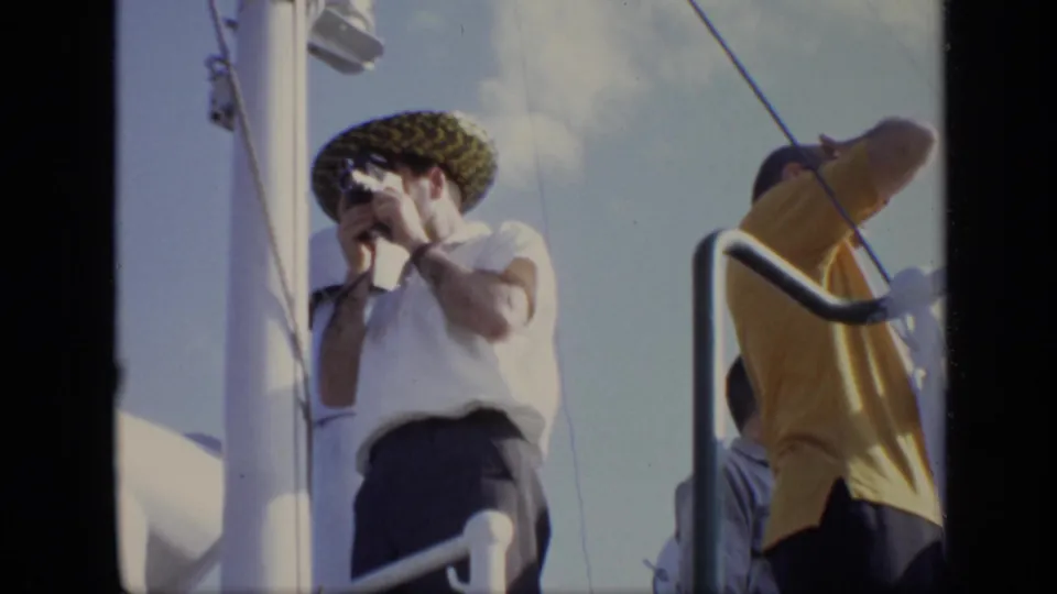 1971 8mm home-movie deck scene with passengers near railings and a man in a patterned hat looking through a handheld camera.