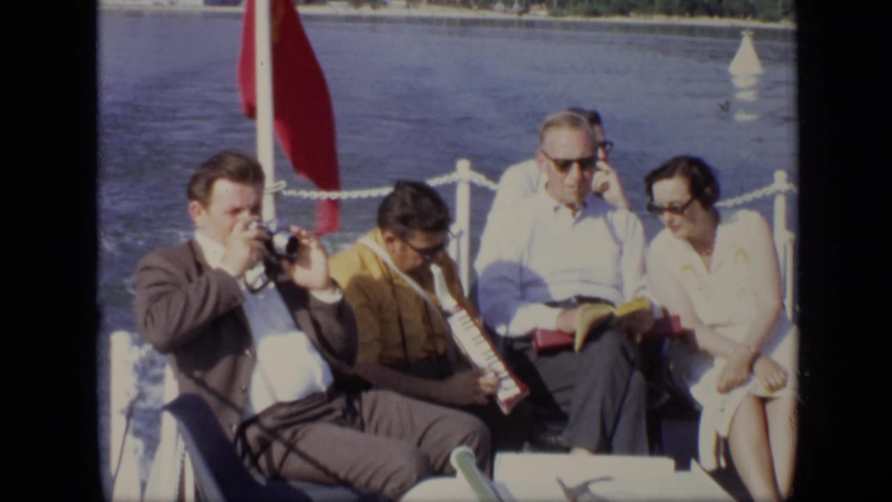 Adults seated on a boat beside water, with one man using a camera and another holding an accordion.