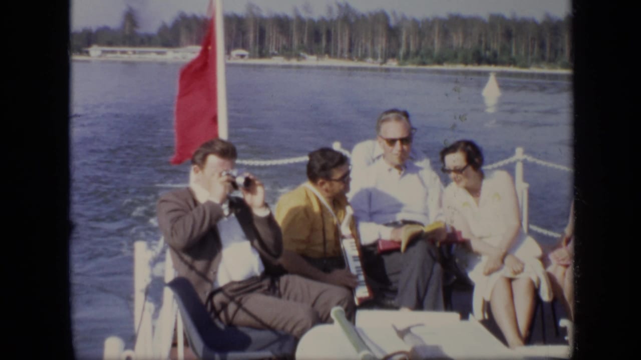 Group of adults seated on a boat with a red flag, water, and shoreline behind them.