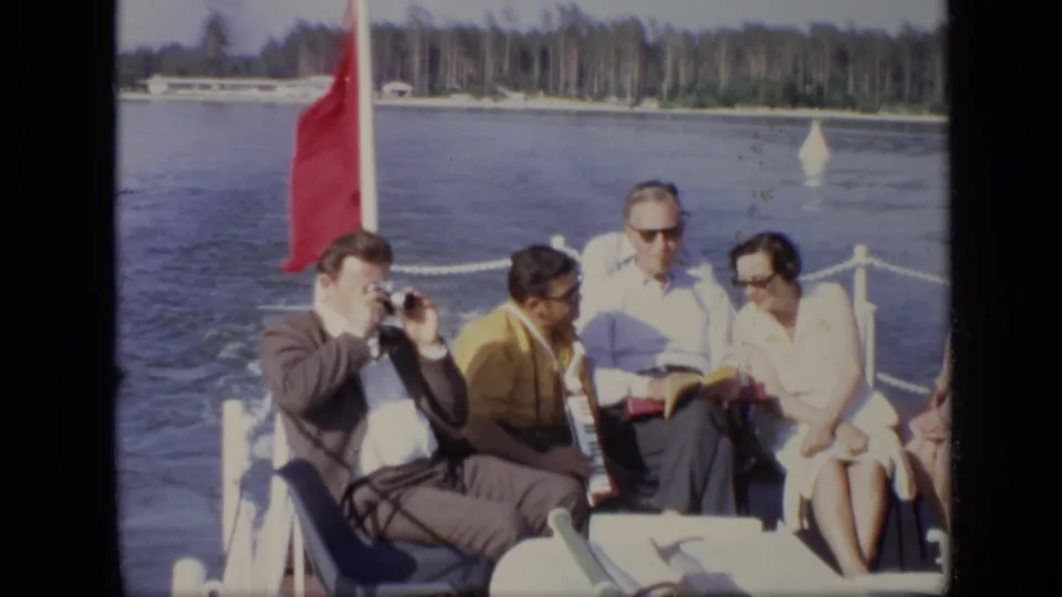 1971 Moscow 8mm frame of adults on a small boat, including a camera user and accordion player, with calm water and trees behind them.