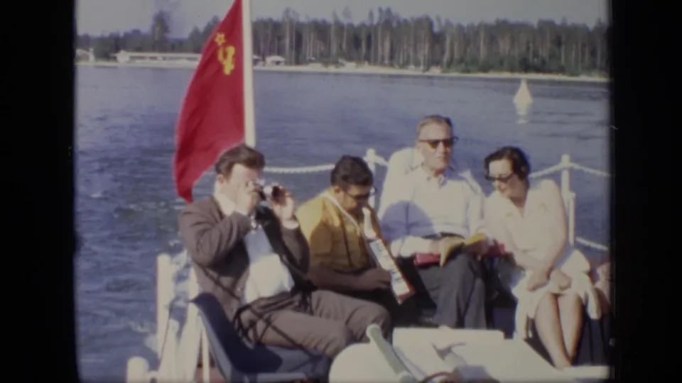 1971 Moscow home-movie view of adults seated close together on a boat, with a red flag, accordion, camera, water, and shoreline visible.