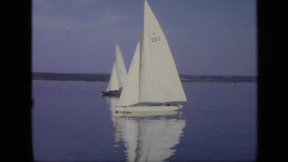 1971 Moscow home-movie frame showing white sailboats gliding across calm lake water under a pale sky.