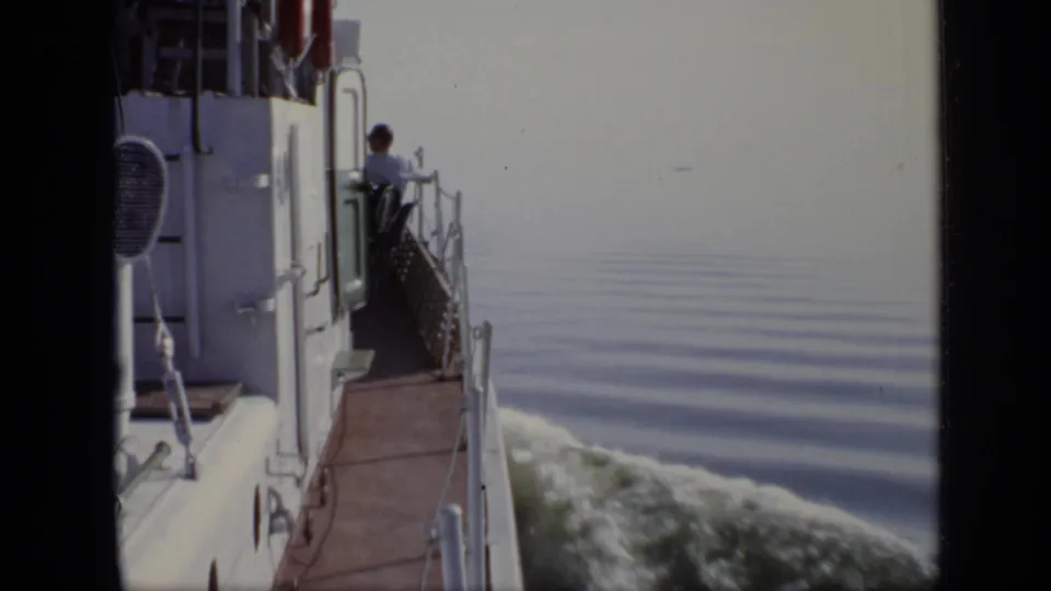 From aboard a moving vessel, a person stands near the deck rail as the boat leaves a wake on calm water.