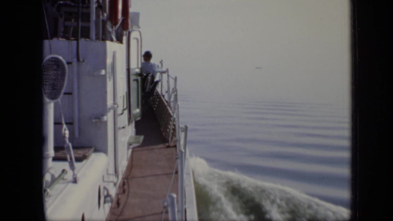 1971 8mm frame showing a white vessel moving across calm water as a man stands near the side railing.