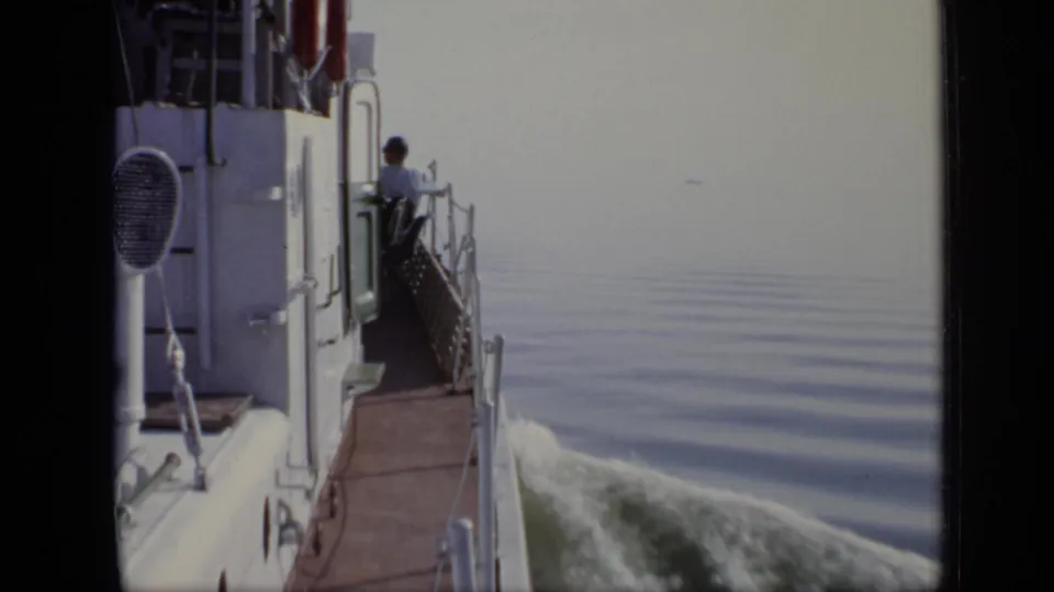1971 8mm frame showing a white vessel moving across calm water as a man stands near the side railing.