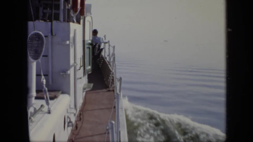 A white ship cuts through calm water, with a man partly visible near the rail in this 1971 home-movie frame.