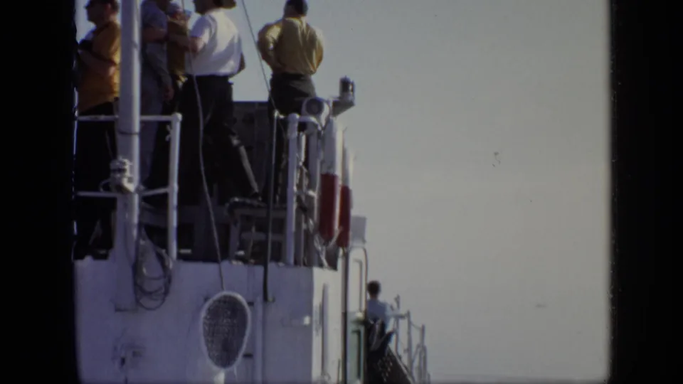 1971 8mm view of men gathered on an upper ship deck, with railings, deck equipment, and bright sky visible.