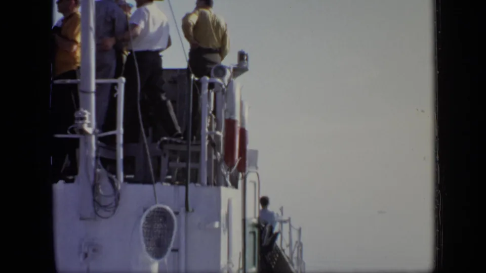 A group of men stands on the ship deck while another figure sits lower near the railing, with open sky behind them.
