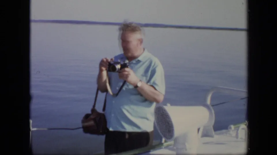 A man in a light blue short-sleeved shirt holds a camera on a boat deck in the supplied 1971 Moscow footage.