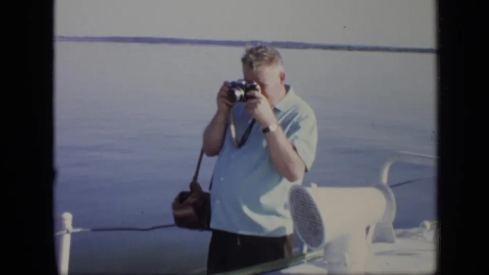 A man raises a still camera to take a photograph on a boat deck in the supplied 1971 Moscow home movie.
