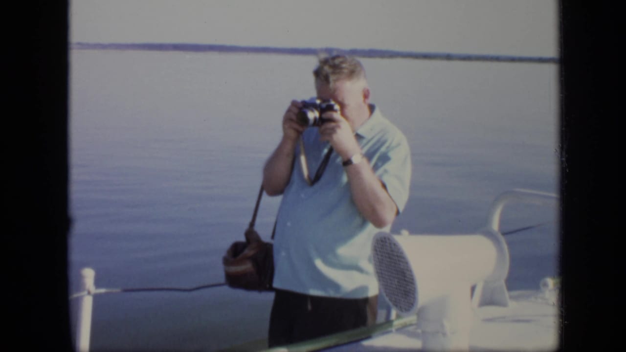 Man in a light blue shirt photographs from a vessel beside calm water.