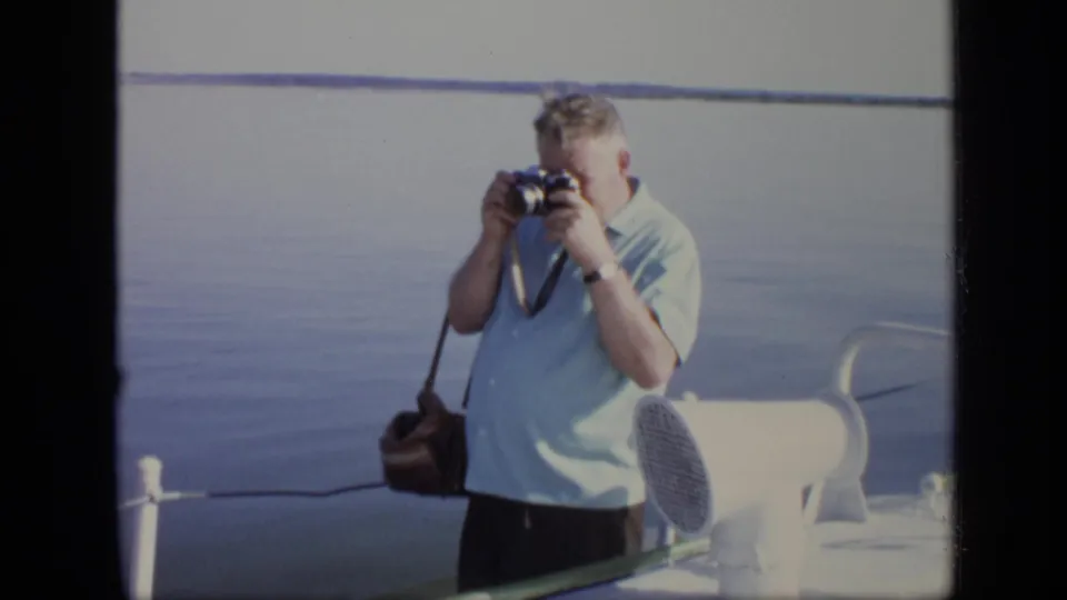 In 1971 Moscow home-movie footage, a man in a light blue shirt raises a camera while standing on a river vessel.