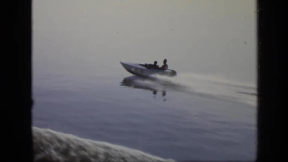 1971 8mm frame of a small motorboat carrying two people across calm water near Moscow, leaving a pale wake.