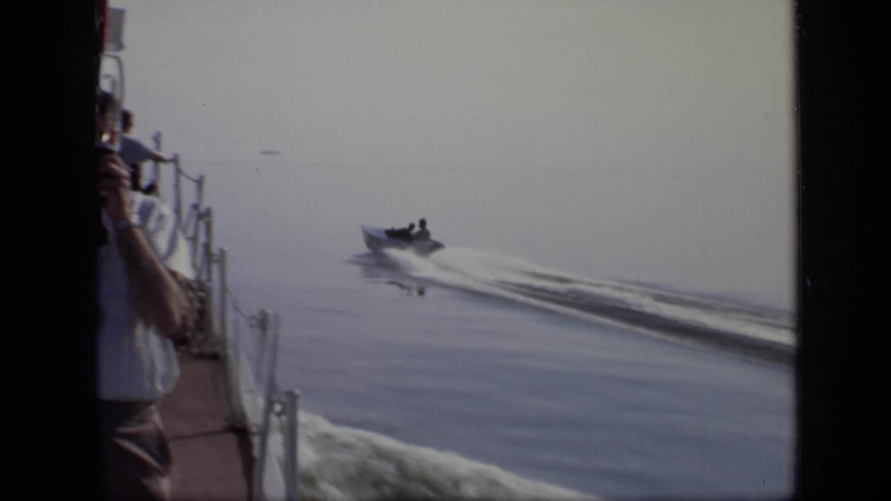Handheld 8mm view from a larger vessel, with a small motorboat crossing the lake and an adult on deck partly visible.