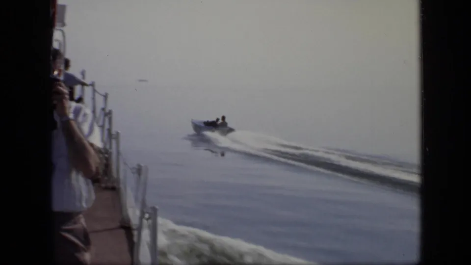 Handheld 8mm view from a larger vessel, with a small motorboat crossing the lake and an adult on deck partly visible.