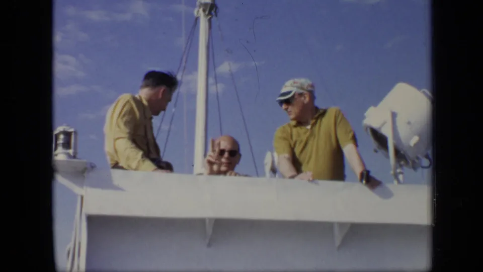 Three adult men on a boat deck in 1971 Moscow home-movie footage, with one wearing sunglasses and another in a yellow shirt and cap.
