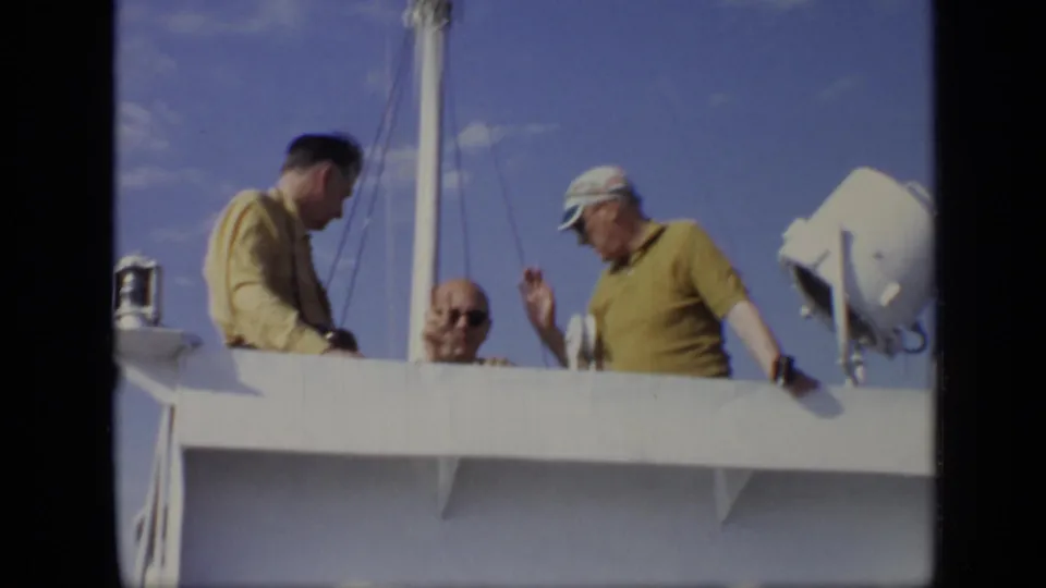 Adult men stand on the deck of a larger boat, with one man in a yellow shirt and cap gesturing under a bright sky.