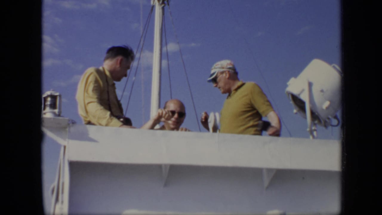 Three adult men on a white boat deck in 1971 Moscow home-movie footage, with mast rigging and sky behind them.