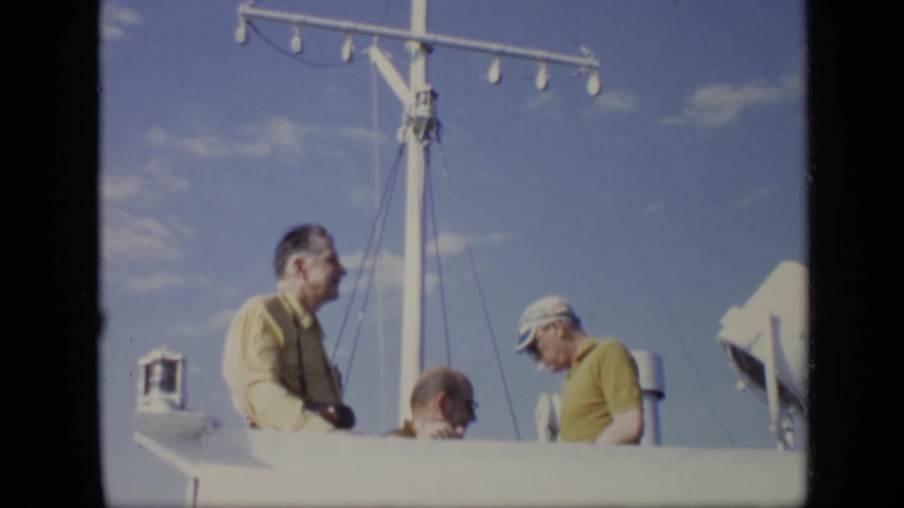 Adult men stand and sit near the mast on a white boat deck, with clear sky and boat equipment visible.