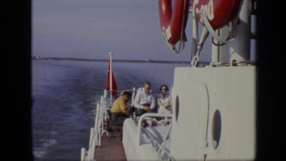 Seated passengers are visible at the stern of a boat on calm water, with a red flag and life preservers above them.