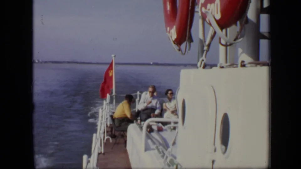 A small group of passengers sits at the stern below red life preservers, with a red flag and water wake behind the boat.