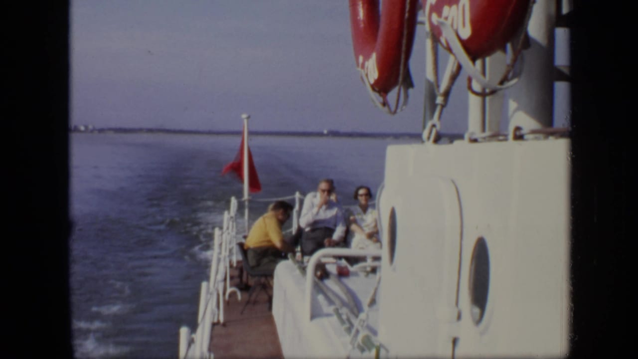 Adults seated on the stern of a white boat with a red flag and water behind them.