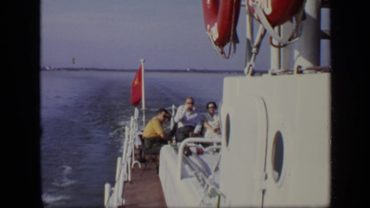 Group of adults seated at the back of a boat near a red flag and white railings.