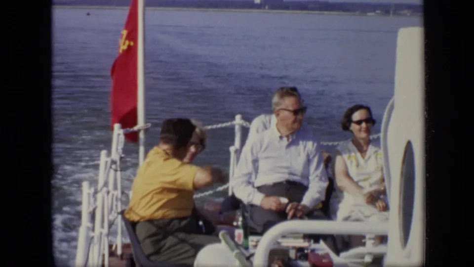1971 Moscow home-movie frame of adults seated on the stern deck beside a red flag while the boat continues across the water.