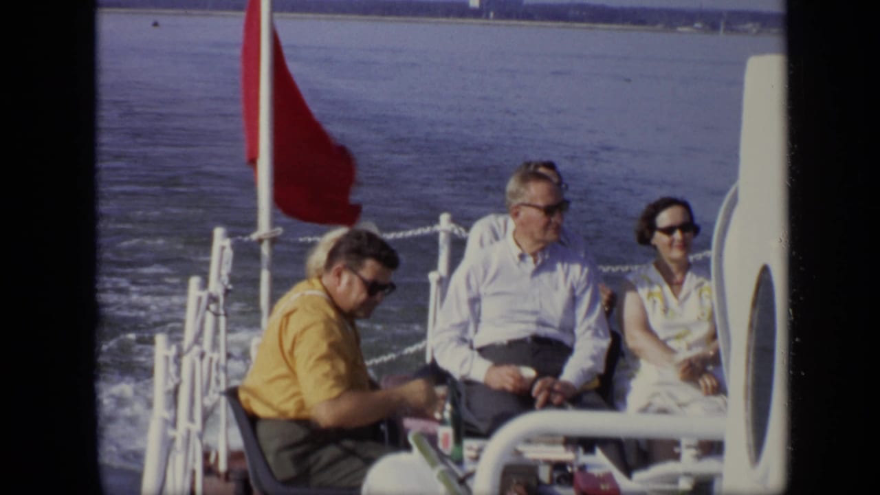 Passengers sit on an open boat deck beside a red flag with river water behind them.
