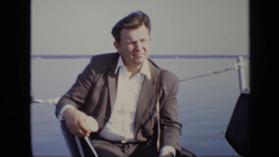 1971 Moscow home-movie frame showing a suited man seated beside white railings on a calm riverboat deck.