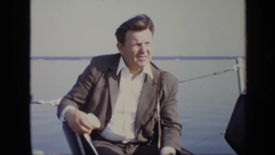 1971 Moscow riverboat frame of a seated man in a brown suit looking across the deck with river and railing behind him.