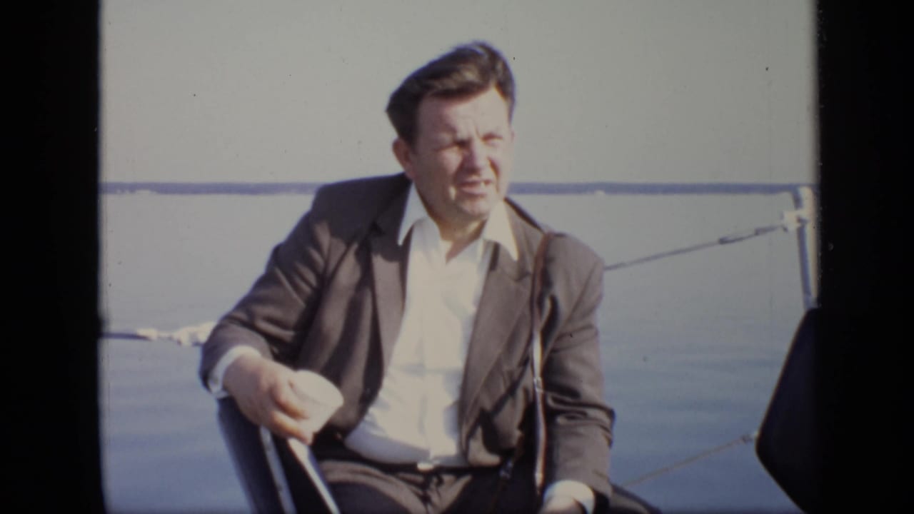Man in a dark suit sits on a boat with water and a chain rail behind him.