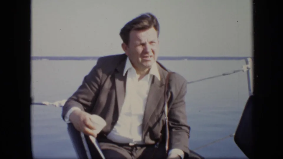 In 1971 Moscow home-movie footage, a seated man in a dark suit and white shirt appears on a boat with water in the background.