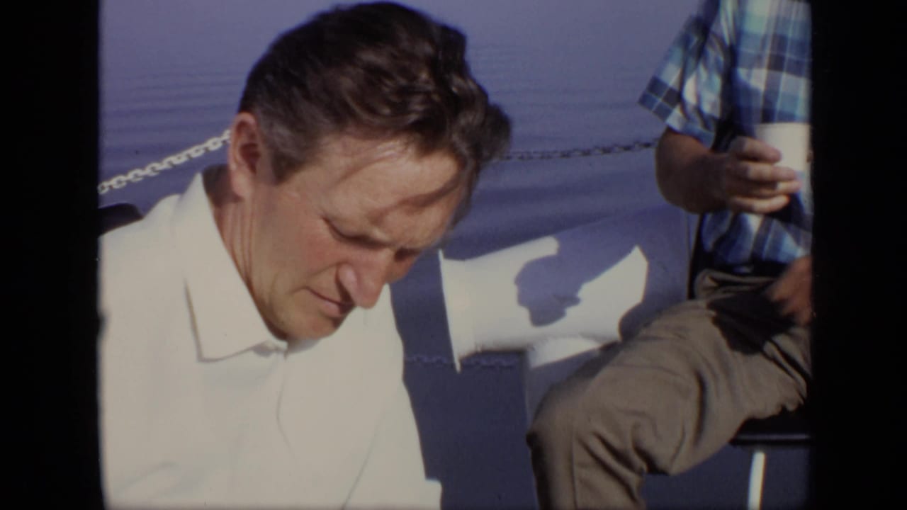 A man in a white shirt looks down on a boat while a second man in a blue plaid shirt holds a cup beside him.