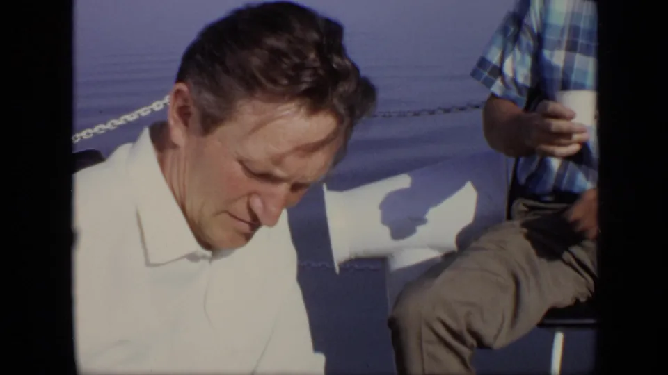 A man in a white shirt looks down on a boat while a second man in a blue plaid shirt holds a cup beside him.