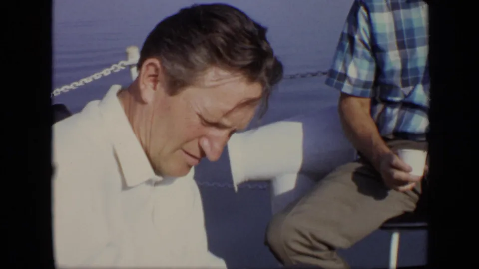Close 8mm frame of a man in a white shirt on a boat, with another seated man in a blue plaid shirt holding a cup nearby.