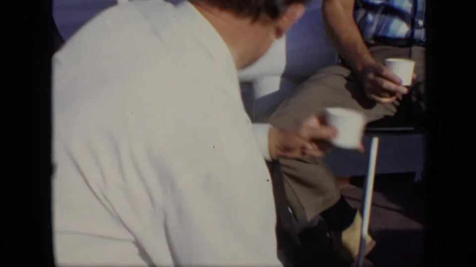 A man in a white shirt turns away from the camera as nearby seated men hold cups on the boat in 1971 Moscow footage.