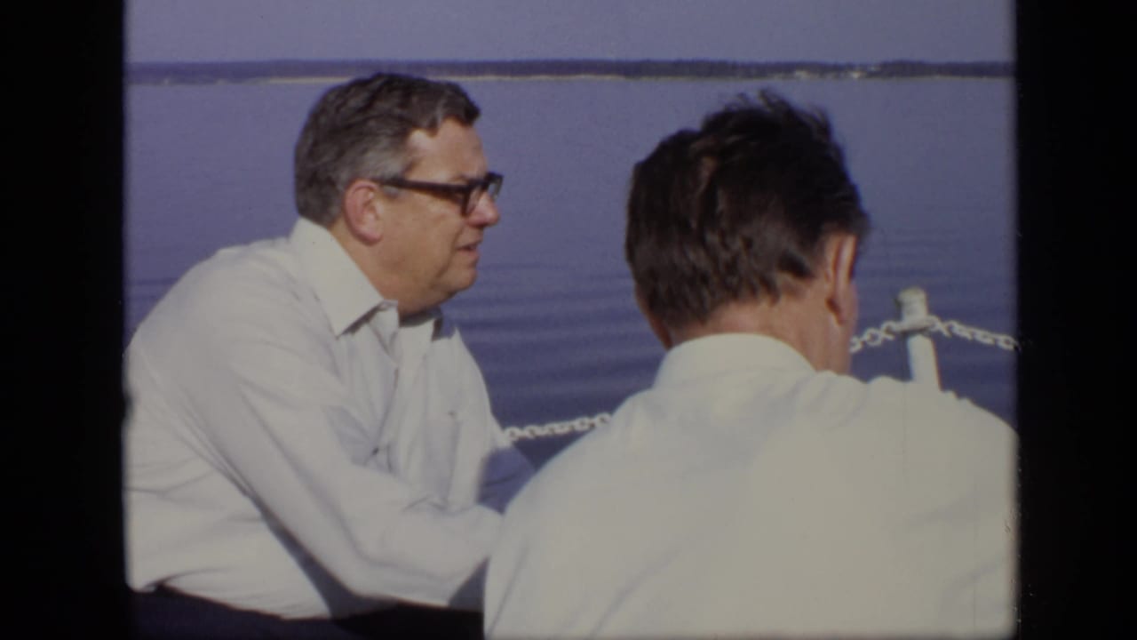 Man with glasses in a white shirt sits near another man with his back to camera by the water.