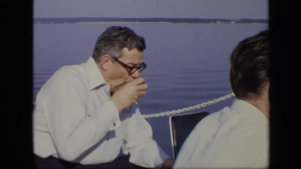1971 Moscow home-movie frame showing a seated man in glasses lifting a cup near other men and a chain railing by the lake.