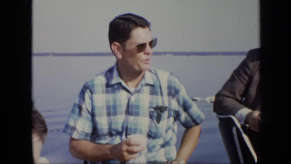 1971 Moscow frame showing a man in a blue plaid shirt and sunglasses seated near calm water, with other adults partly visible.