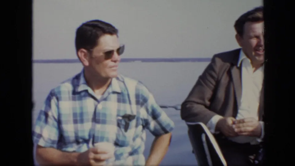 Two adult men sit side by side on a boat in 1971 Moscow home-movie footage, one in sunglasses and a plaid shirt.