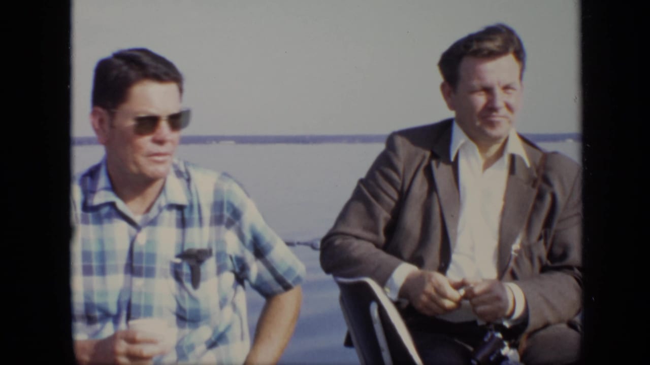 In the 1971 Moscow boat scene, the man in a suit jacket sits beside a man in sunglasses and appears to handle a camera.